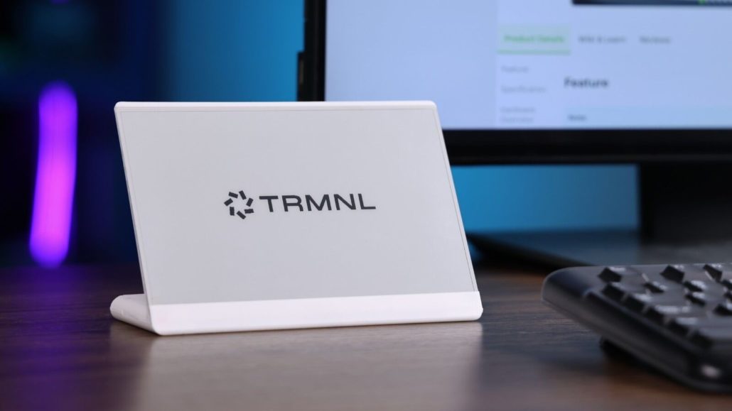A white TRMNL epaper sits on a desk in front of a keyboard and a computer monitor.